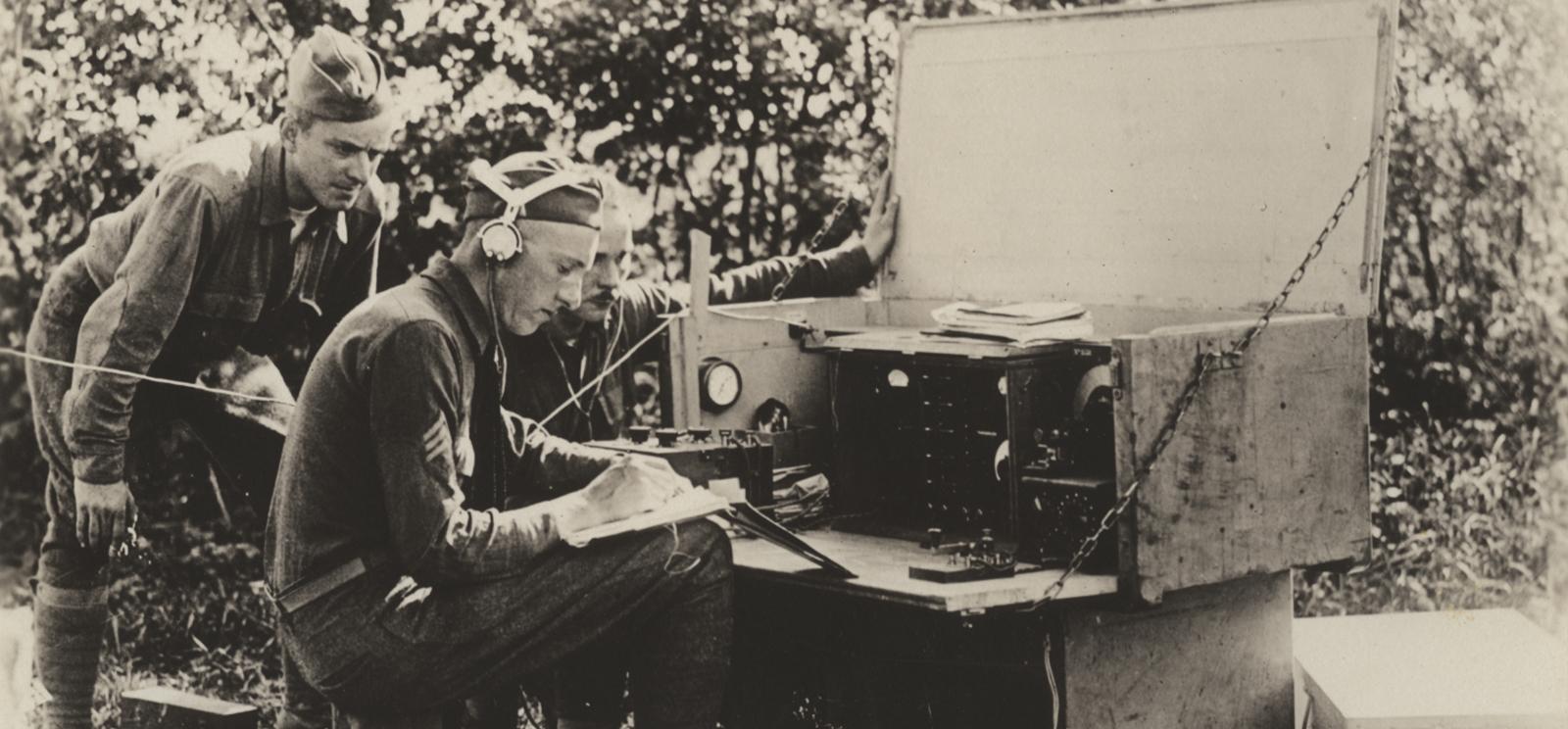 Sepia photograph of a WWI soldier seated at a field radio station wearing headphones and writing something on paper. Two soldiers lean over his shoulder.