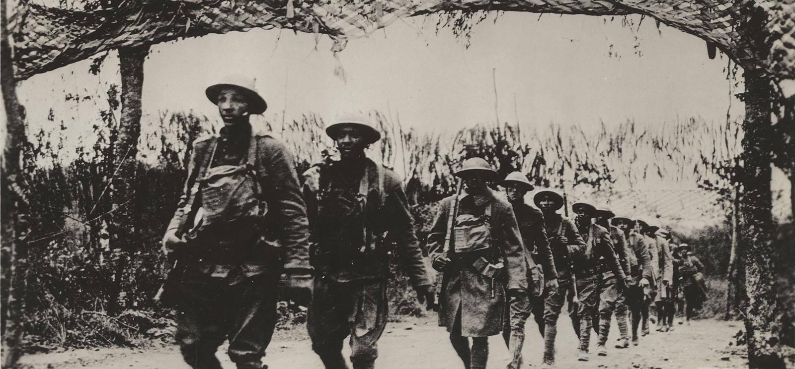 Black and white photo of a line of WWI-era Black soldiers walking in a line down a path in a forest.