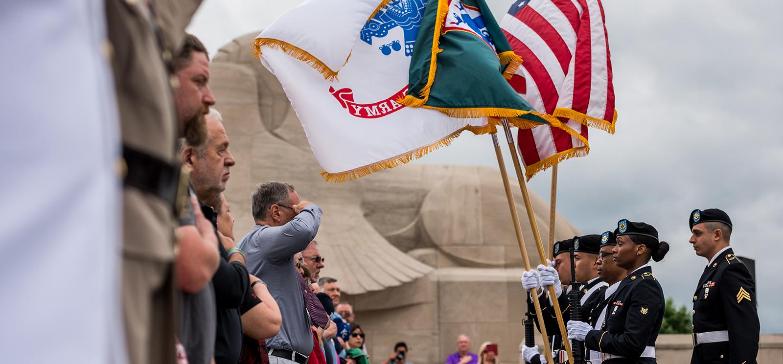 Color guard bearing flags facing a row of civilians either saluting or standing respectfully