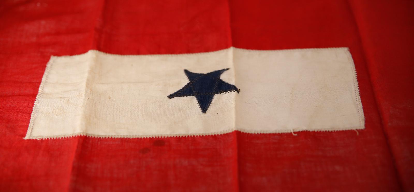 Modern photograph of a WWI-era cloth flag. A red border surrounds a white/cream rectangle with a single dark blue star embroidered in the middle.