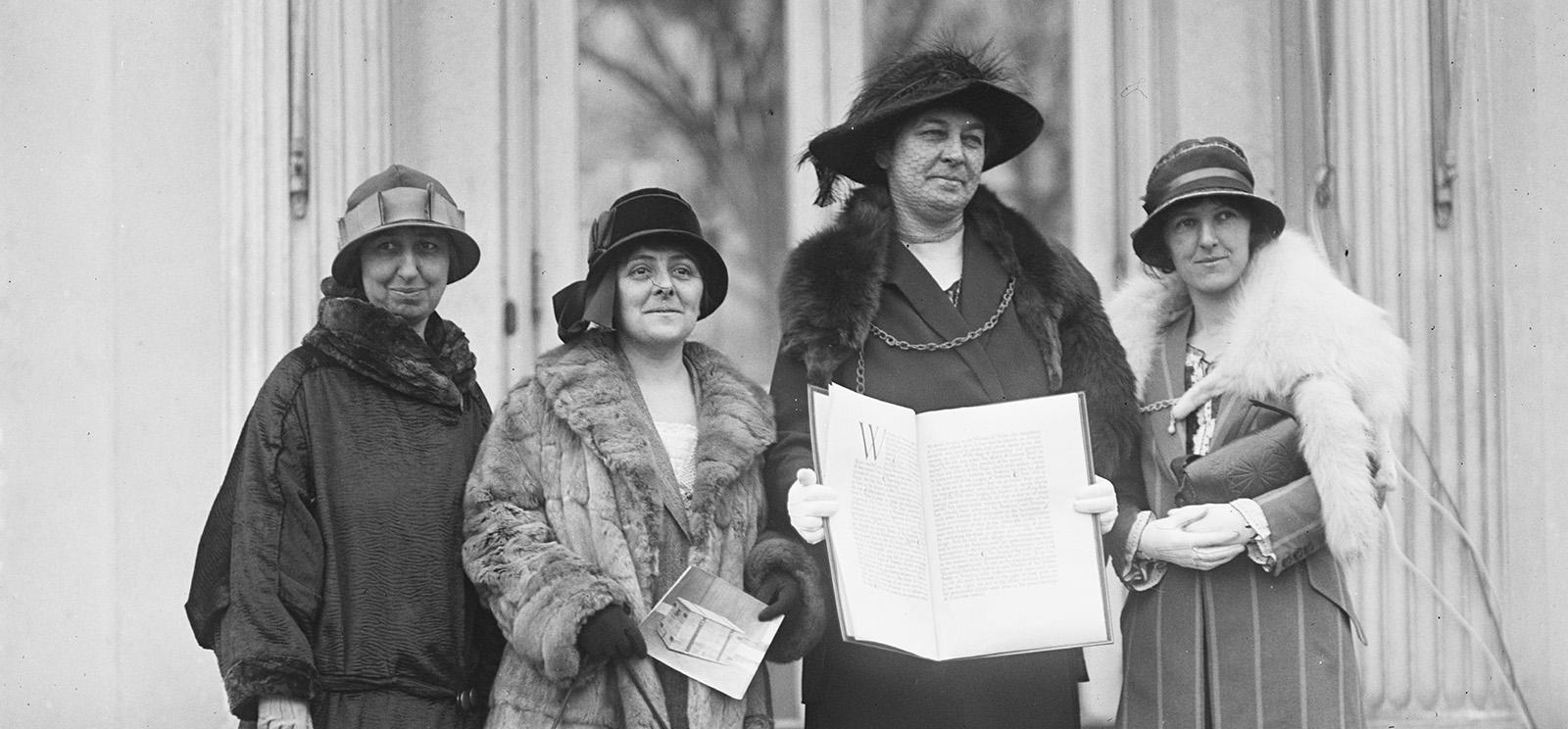 Black and white photograph of four white women dressed in fine traveling dresses, coats and hats posing outside with one woman holding open a large folio-like object with writing on the pages.