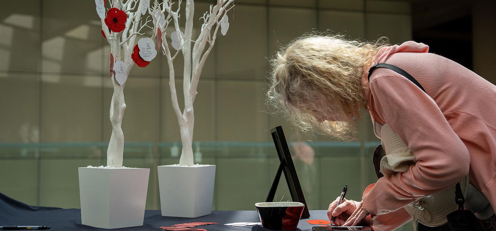 Modern photograph of a table on the Glass Bridge. Several small artificial trees painted white, with red paper poppies hung on their branches, stand on the table. A white woman with blonde hair is bent over the table writing something on a paper poppy with a pen.