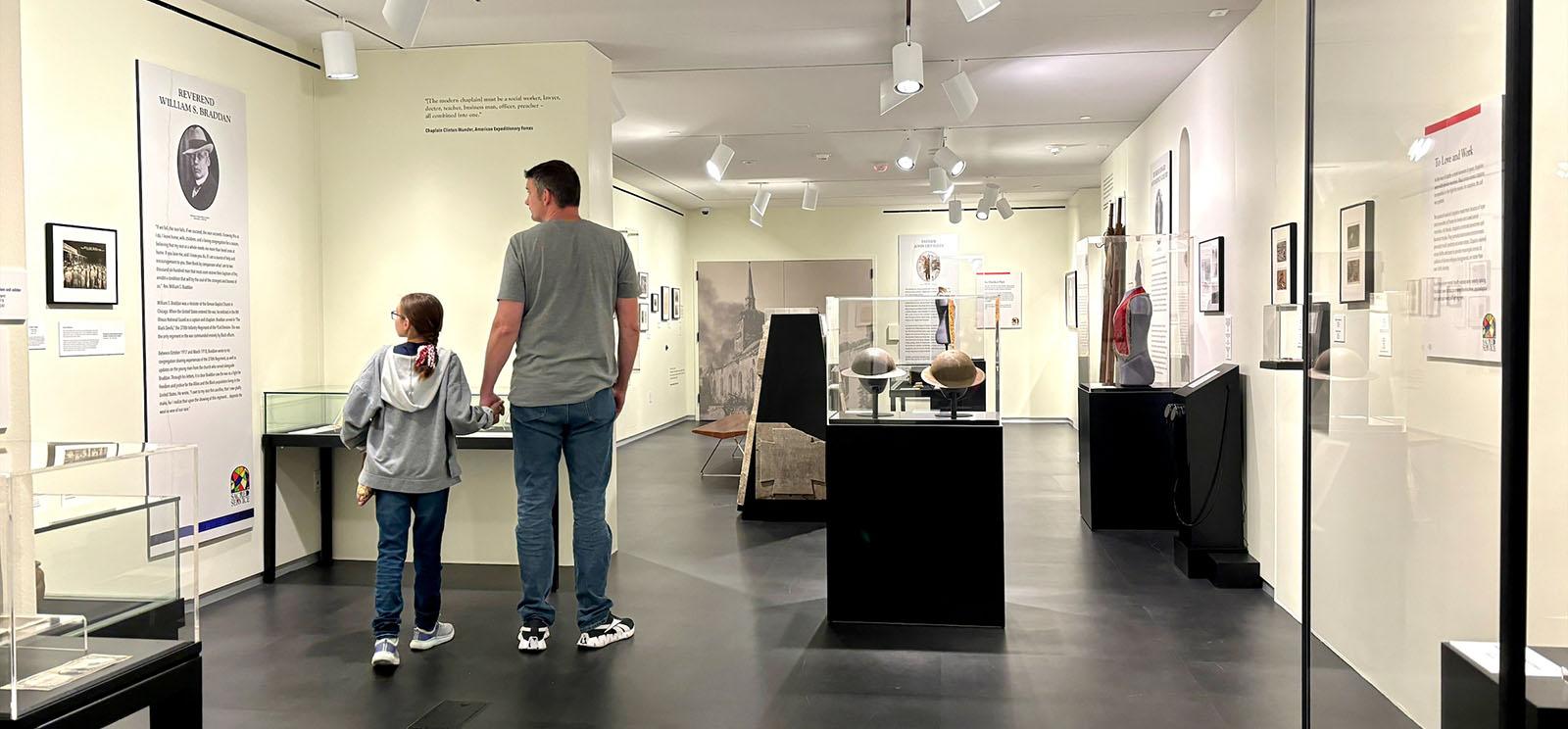 Modern photograph wide shot of a museum gallery. The walls are painted creamy white and the lighting is bright on glass cases displaying artifacts like WWI helmets and religious vestments. Two people are looking at a wall panel of text, facing away from the viewer: a tall white man dressed casually in jeans and t-shirt, holding hands with a young white girl in jeans and a hoodie.