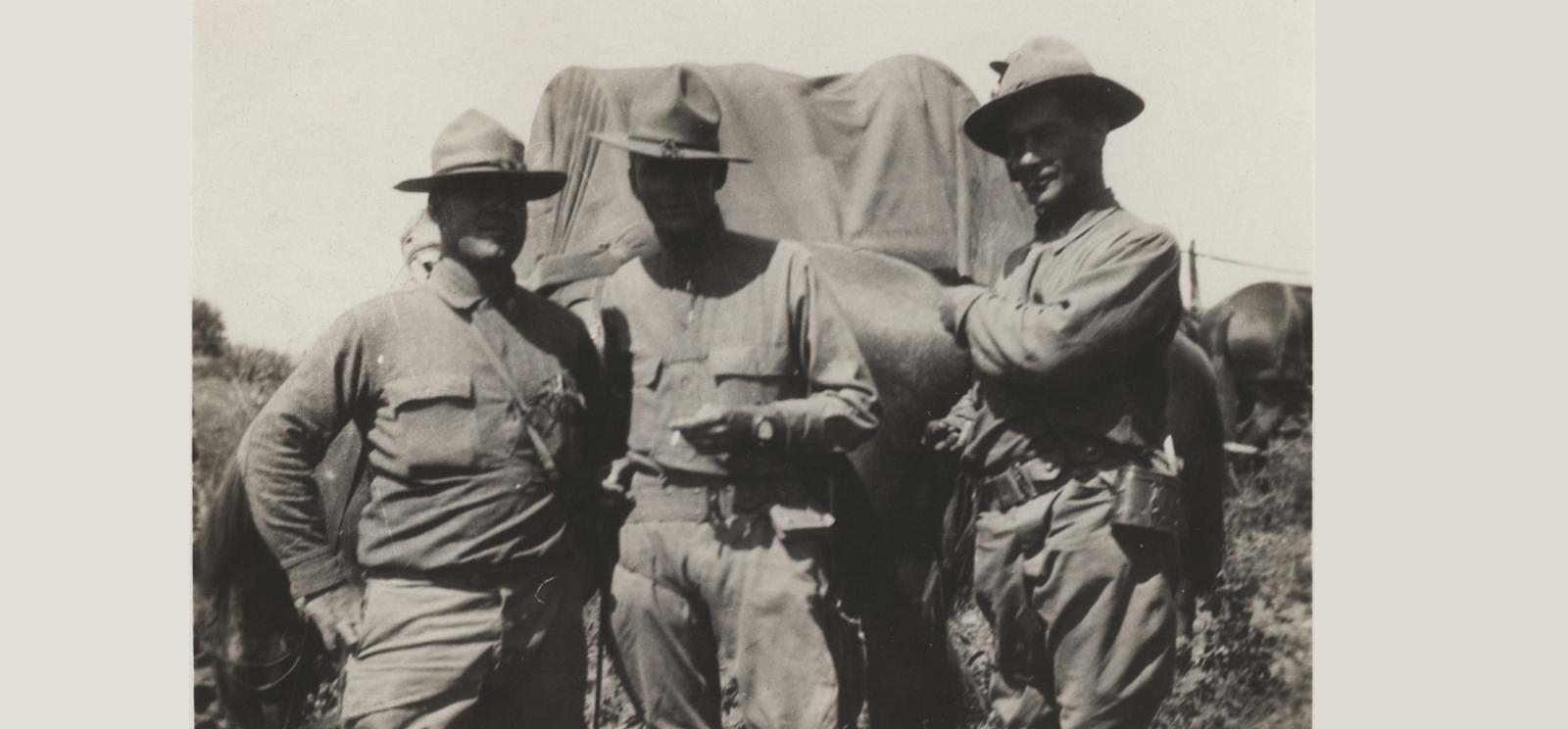 Black and white photograph of three unidentified soldiers posing for a picture. Center man is smoking a cigarette. In background is a covered wagon and horses. 