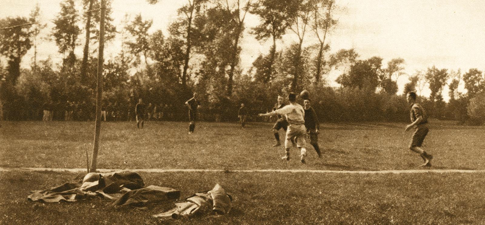 Sepia photo of several WWI soldiers on a field playing football (soccer). In the foreground, discarded equipment and clothing lie under a tree.