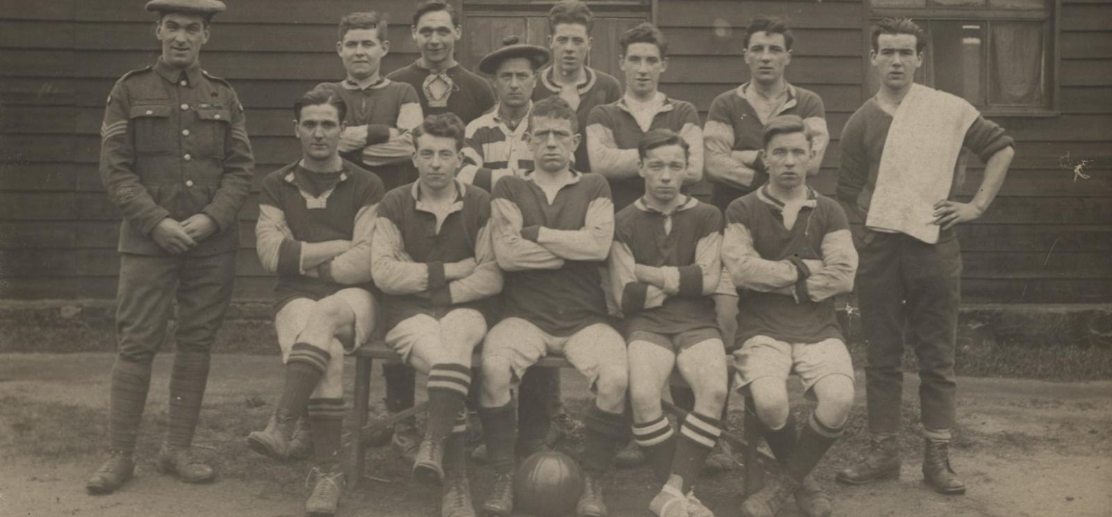 Black and white photograph of a group of men. The first row is sitting, and the rest are all standing, with the majority in football uniforms. One man stands to the side carrying a towel and another stands on the opposite side in uniform. There is a soccer ball at the front between the legs of one of the men.