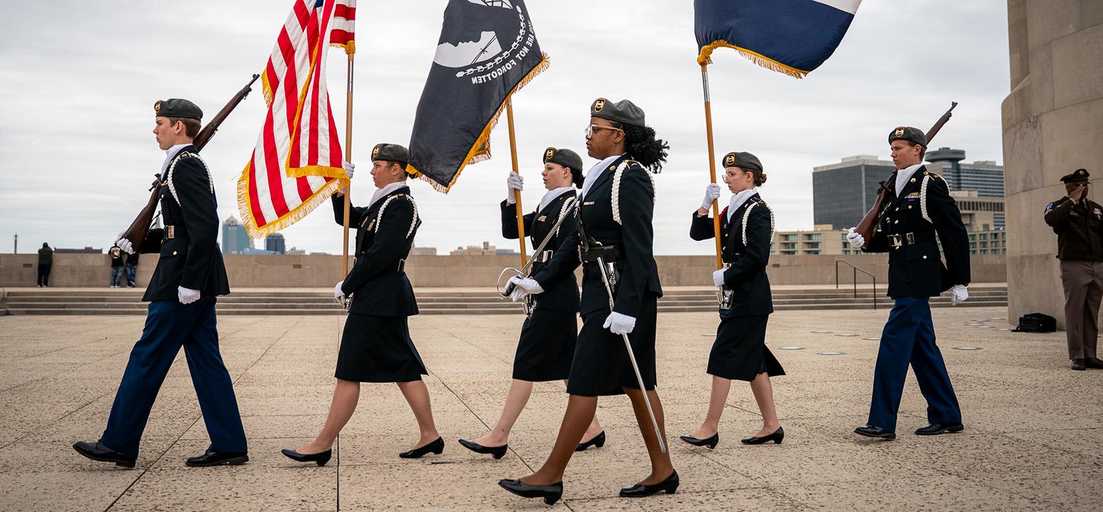Modern photo of six young people in military uniform marching in a line carrying flags, rifles or swords
