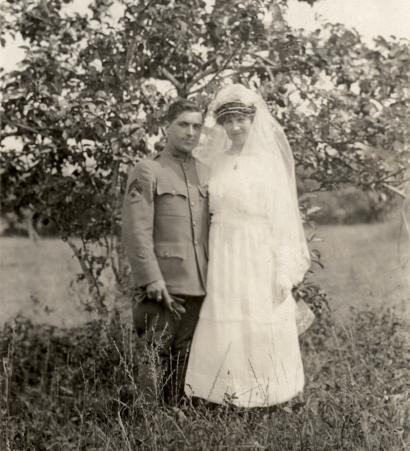 Photographie en noir et blanc d'une femme blanche et d'un homme blanc debout devant un arbre. La femme porte une robe de mariée blanche et un voile et l'homme est en uniforme militaire américain.