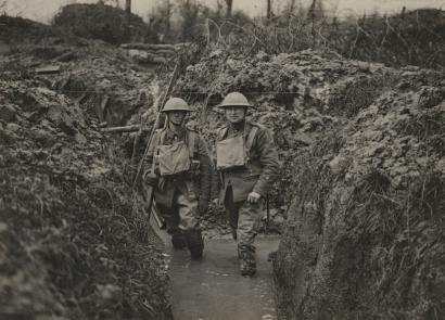 Black and white photograph of two men in military combat gear and steel helmets walking toward the viewer in a trench. They are up to their ankles in water.