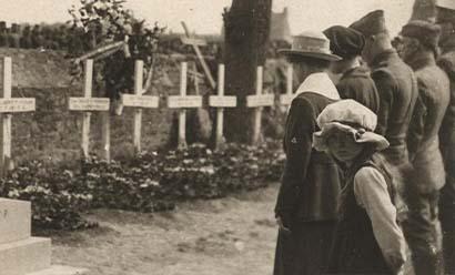 Sepia photograph of a row of people standing in front of a row of crosses. In the foreground a young girl in a large hat turns to look at the viewer.