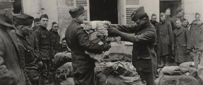 Photograph of two soldiers amidst other soldiers standing around them. One of them has an armful of parcels while the other soldier hands him more.