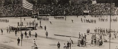 Black and white photograph. View of a wide open sports field in a stadium filled with spectators. Different track and field events are taking place in different spots on the field. 