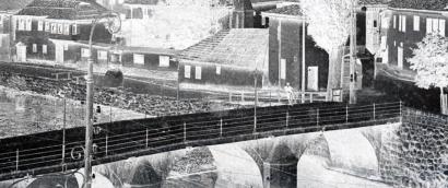 Blackand white photograph with colors inverted so it looks like a photo negative. The image shows a long-distance view of a bridge over a river. The river edge is lined with low houses. There is one person leaning against the railing of the bridge.