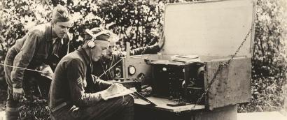Black and white photograph of three soldiers gathered around a large field radio.
