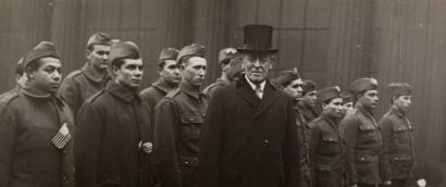 Black and white photo of Woodrow Wilson in a top hat standing in front of a line of soldiers in military uniform.