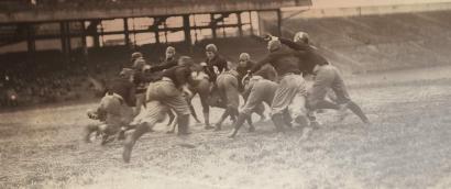 Black and white action shot of a group of North American football players mid-play.