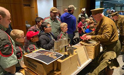 Modern photograph of children gathered in front of a long table filled with WWI-era artifacts and reproduction. A man dressed in WWI-era uniform demonstrates with one of the artifacts in his hands.