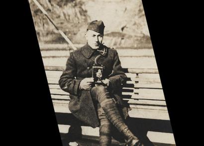 Black and white photograph of a man in military uniform sitting down on a bench. He holds a WWI-era personal camera in his lap, aimed at the viewer.
