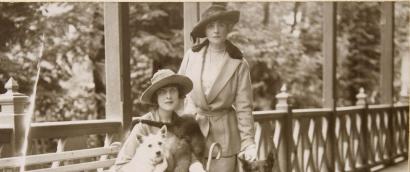 Sepia photograph of two women dressed in walking suits and hats on a building's porch. One is seated with a small dog in her lap.