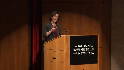 Modern photograph of a white brunette woman standing at the National WWI Museum and Memorial podium on the auditorium stage. She is speaking and gesturing energetically.