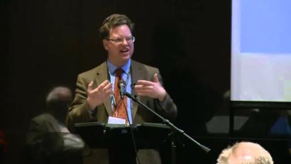 Video still of a white man in a brown suit giving a presentation to an audience.