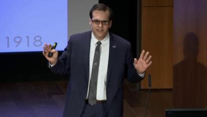 Video still of a white man in aa suit giving a presentation on the Museum auditorium stage.