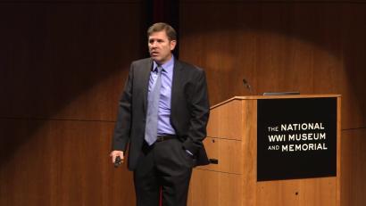 Video still of a white man in a suit giving a presentation on the Museum auditorium stage.
