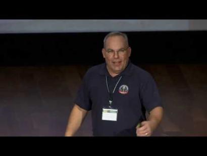 Video still of a middle-aged white man wearing a blue polo shirt giving a presentation on the Museum auditorium stage.