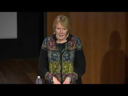 Video still of a white woman in a colorful top speaking at a podium on the Museum auditorium stage.