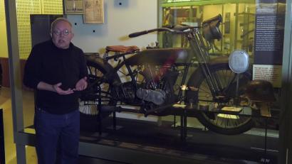 Video still of an older white man in a black shirt, facing the viewer, standing in front of a museum display of a WWI-era motorcycle.