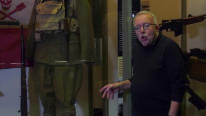 Video still of an older white man facing the viewer, standing in front of a museum display of a WWI-era military combat uniform.