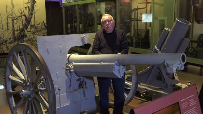 Video still of an older white man facing the viewer, standing by a museum display of a WWI-era trench gun.