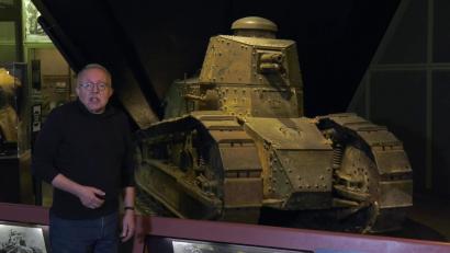 Video still of an older white man facing the viewer, standing in front of a museum display of a small WWI-era tank.