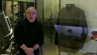 Video still of an older white man facing the viewer, standing in front of a museum display of a military uniform.