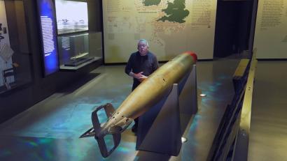 Video still of an older white man facing the viewer, standing by a museum display of a WWI-era torpedo.