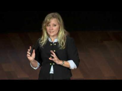 Video still of a blonde woman giving a presentation on the auditorium stage.