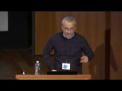 Video still of an older man in a sweater giving a presentation at the podium on the Museum auditorium stage.