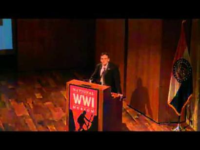 Video still of a man in a suit standing at a podium on the Museum auditorium stage.