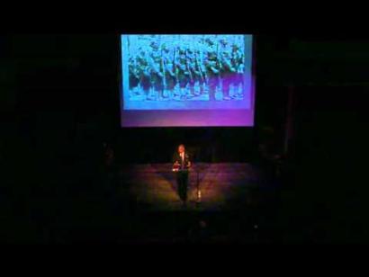 Video still of a Black man at the podium giving a presentation on the Museum auditorium stage.