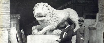 Black and white photograph of a man in a suit stsanding in front of a large stone statue of a lion.