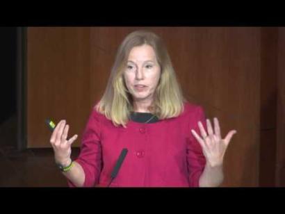 Video still of a blonde woman in a pink top giving a presentation on the Museum auditorium stage.