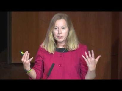 Video still of a blonde woman in a pink top giving a presentation on the Museum auditorium stage.