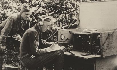 Sepia photograph of a WWI soldier seated at a field radio station wearing headphones and writing something on paper. Two soldiers lean over his shoulder.