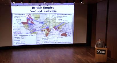Screencap from video of speaker at podium on a stage, with powerpoint presentation on screen behind with text reading British Empire,confused leadership and a map