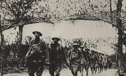 Black and white photo of a line of WWI-era Black soldiers walking in a line down a path in a forest.