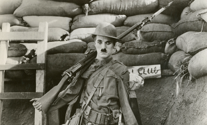 Black and white promotional still of Charlie Chaplin dressed in WWI uniform with a rifle over his shoulder standing in a trench.