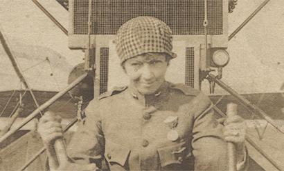 Sepia photograph of a white woman in an aviation uniform sitting in the cockpit of an WWI-era airplane