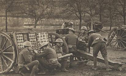 Sepia photograph of six young men wearing WWI uniforms and helmets gathered around a field artillery gun in a grassy area bounded by trees.