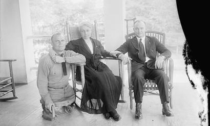 Black and white photo of three people posed for a group photo. Left to right: a white man in military uniform with both legs amputated nearly at the hip, resting on a small platform with wheels. A white woman in a dark-colored dress sitting in a rocking chair. A white man wearing a suit sitting in a rocking chair with a crutch leaning on a pillar behind him. The woman has her arm around the shoulders of the man on the left and her hand on the arm of the man on the right.