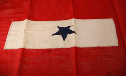 Modern photograph of a WWI-era cloth flag. A red border surrounds a white/cream rectangle with a single dark blue star embroidered in the middle.